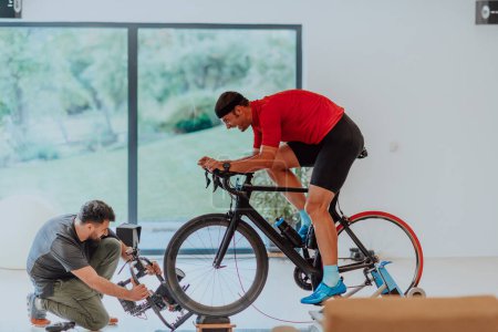 Foto de Un camarógrafo filmando a un atleta montando una bicicleta de triatlón en una máquina de simulación en una sala de estar moderna. Formación en condiciones pandémicas - Imagen libre de derechos