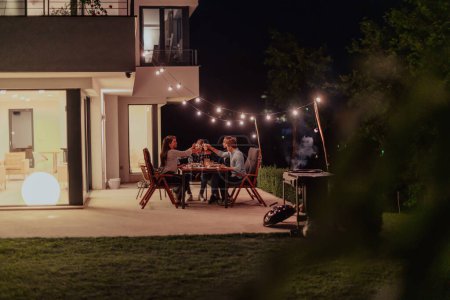 Téléchargez les photos : Un groupe de jeunes gens divers dînant sur la terrasse d'une maison moderne le soir. Du plaisir pour les amis et la famille. Célébration des fêtes, mariages avec barbecue - en image libre de droit