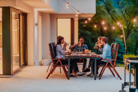 Téléchargez les photos : Un groupe de jeunes gens divers dînant sur la terrasse d'une maison moderne le soir. Du plaisir pour les amis et la famille. Célébration des fêtes, mariages avec barbecue - en image libre de droit