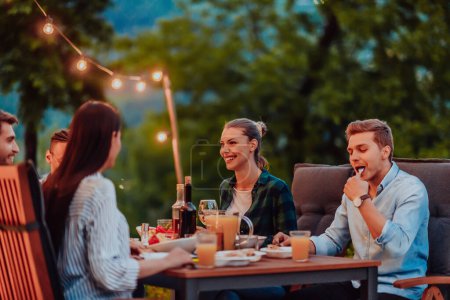 Photo for A group of young diverse people having dinner on the terrace of a modern house in the evening. Fun for friends and family. Celebration of holidays, weddings with barbecue - Royalty Free Image