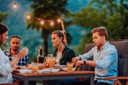 Foto de Un grupo de jóvenes diversos cenando en la terraza de una casa moderna por la noche. Diversión para amigos y familiares. Celebración de fiestas, bodas con barbacoa - Imagen libre de derechos