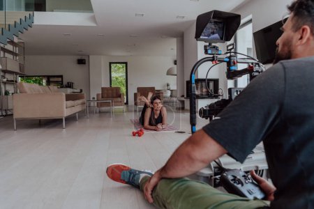 Photo for Behind the scene. Photo of a cameraman recording a woman doing yoga exercises at an online training while using and watching a laptop. - Royalty Free Image