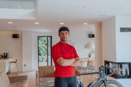 Foto de Foto de un determinado triatleta de pie en una gran sala de estar moderna, detrás de él está su bicicleta de entrenamiento. - Imagen libre de derechos