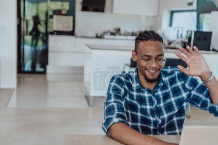 Foto de Hombre afroamericano en gafas sentado en una mesa en una sala de estar moderna, utilizando una computadora portátil para chat de video de negocios, conversación con amigos y entretenimiento. - Imagen libre de derechos