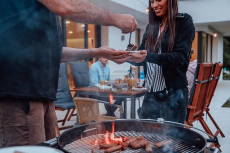 Photo for A group of friends and family barbecue together in the evening on the terrace in front of a large modern house. - Royalty Free Image