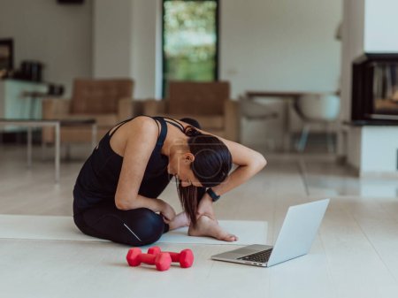 Téléchargez les photos : Une jeune femme en vêtements de sport est assise dans le salon et se prépare à la formation en ligne tout en utilisant un ordinateur portable. - en image libre de droit