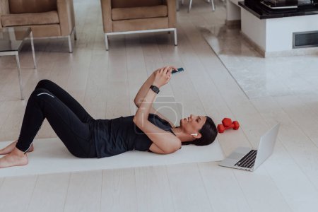 Téléchargez les photos : Sportswoman utilisant un smartphone tout en se reposant et couché sur le sol de la maison moderne. Photo de haute qualité - en image libre de droit