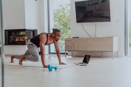Foto de Entrenamiento en casa. Hombre deportivo haciendo el entrenamiento mientras ve un tutorial en línea en un ordenador portátil, haciendo ejercicio en la sala de estar, espacio libre. - Imagen libre de derechos