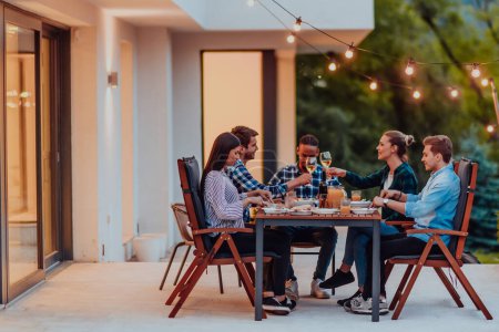 Foto de Un grupo de jóvenes diversos cenando en la terraza de una casa moderna por la noche. Diversión para amigos y familiares. Celebración de fiestas, bodas con barbacoa - Imagen libre de derechos