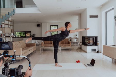 Photo for Behind the scene. Photo of a cameraman recording a woman doing yoga exercises at an online training while using and watching a laptop. - Royalty Free Image