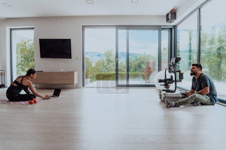 Foto de Detrás de escena. Foto de un camarógrafo grabando a una mujer haciendo ejercicios de yoga en un entrenamiento en línea mientras usa y mira una computadora portátil. - Imagen libre de derechos