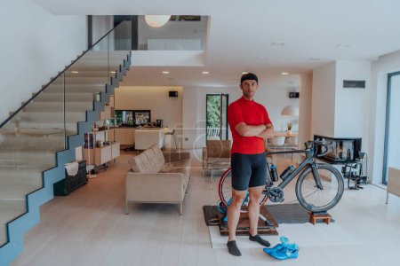 Foto de Foto de un determinado triatleta de pie en una gran sala de estar moderna, detrás de él está su bicicleta de entrenamiento. - Imagen libre de derechos
