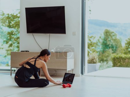 Photo for A young woman in sportswear is sitting in the living room and preparing for online training while using a laptop. - Royalty Free Image