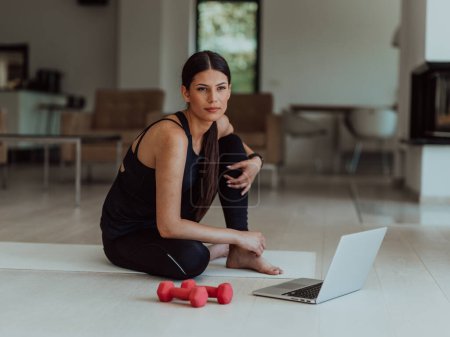 Téléchargez les photos : Une jeune femme en vêtements de sport est assise dans le salon et se prépare à la formation en ligne tout en utilisant un ordinateur portable. - en image libre de droit
