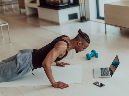Foto de Joven hombre afroamericano trabajando flexiones en la sala de estar mientras ve entrenamiento en línea en el ordenador portátil. - Imagen libre de derechos