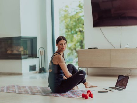Foto de Una joven en ropa deportiva está sentada en la sala de estar y preparándose para el entrenamiento en línea mientras usa una computadora portátil. - Imagen libre de derechos
