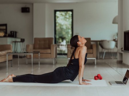Téléchargez les photos : Jeune belle femme faisant de l'exercice, de l'étirement et pratiquant le yoga avec un entraîneur par vidéoconférence dans une maison ensoleillée lumineuse. Mode de vie sain, bien-être et pleine conscience Concept. - en image libre de droit