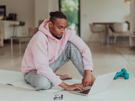 Foto de Hombre afroamericano en gafas sentado en una mesa en una sala de estar moderna, utilizando una computadora portátil para chat de video de negocios, conversación con amigos y entretenimiento. - Imagen libre de derechos