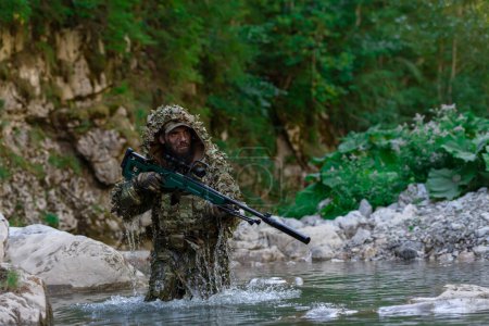 Foto de Un militar o un jugador de airsoft con un traje de camuflaje que se cuela en el río y apunta desde un rifle de francotirador hacia un lado o para apuntar - Imagen libre de derechos