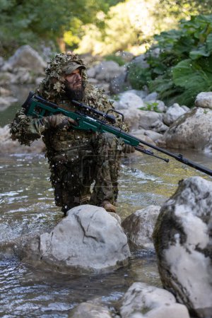 Foto de Un militar o un jugador de airsoft con un traje de camuflaje que se cuela en el río y apunta desde un rifle de francotirador hacia un lado o para apuntar - Imagen libre de derechos