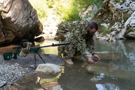 Photo for Soldier in a camouflage suit uniform drinking fresh water from the river. Military sniper rifle on the side - Royalty Free Image