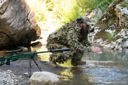 Foto de Soldado en un traje de camuflaje uniforme bebiendo agua dulce del río. rifle de francotirador militar en el lateral - Imagen libre de derechos
