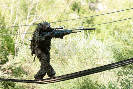 Photo for A military man or airsoft player in a camouflage suit sneaking the rope bridge and aims from a sniper rifle to the side or to target. - Royalty Free Image
