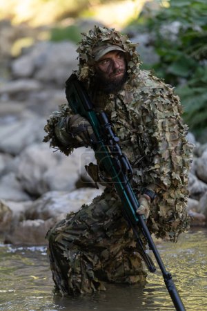 Téléchargez les photos : Un militaire ou un joueur de airsoft en costume de camouflage qui se faufile dans la rivière et vise à tirer d'un fusil de sniper sur le côté ou à cibler - en image libre de droit