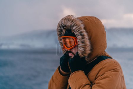 Foto de Foto de un hombre en una zona nevada fría con una chaqueta gruesa de invierno marrón, gafas de nieve y guantes. La vida en las regiones frías del país - Imagen libre de derechos