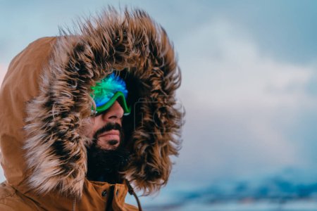 Photo for Headshot photo of a man in a cold snowy area wearing a thick brown winter jacket, snow goggles and gloves. Life in cold regions of the country - Royalty Free Image