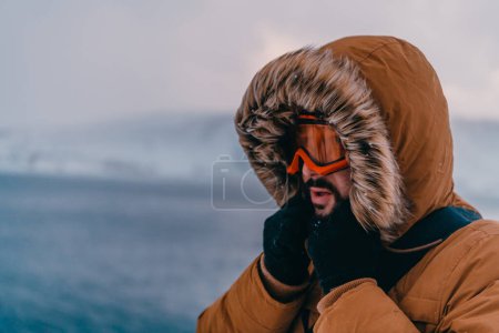 Foto de Foto de un hombre en una zona nevada fría con una chaqueta gruesa de invierno marrón, gafas de nieve y guantes. La vida en las regiones frías del país - Imagen libre de derechos