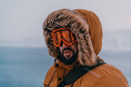 Photo for Headshot photo of a man in a cold snowy area wearing a thick brown winter jacket, snow goggles and gloves. Life in cold regions of the country - Royalty Free Image