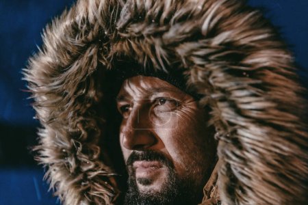Foto de Foto de un hombre en una fría zona nevada con una gruesa chaqueta de invierno marrón, gafas de nieve y guantes en una fría noche escandinava. La vida en las regiones frías del país - Imagen libre de derechos