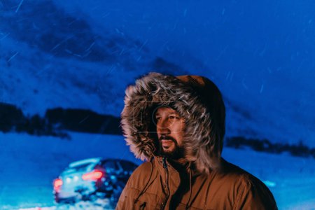 Foto de Foto de un hombre en una fría zona nevada con una gruesa chaqueta de invierno marrón, gafas de nieve y guantes en una fría noche escandinava. La vida en las regiones frías del país - Imagen libre de derechos