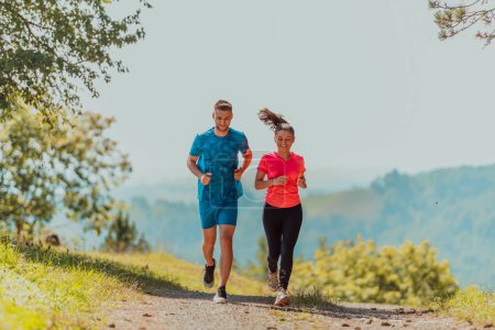 Foto de Couple enjoying in a healthy lifestyle while jogging on a country road through the beautiful sunny forest, exercise and fitness concept. - Imagen libre de derechos