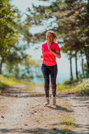 Téléchargez les photos : Jeune femme heureuse jouissant d'un mode de vie sain tout en faisant du jogging sur une route de campagne à travers la belle forêt ensoleillée, concept d'exercice et de remise en forme
. - en image libre de droit