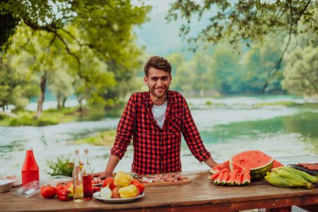 Photo for A man preparing a delicious dinner for his friends who are having fun by the river in nature. - Royalty Free Image