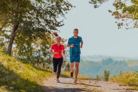 Foto de Couple enjoying in a healthy lifestyle while jogging on a country road through the beautiful sunny forest, exercise and fitness concept. - Imagen libre de derechos