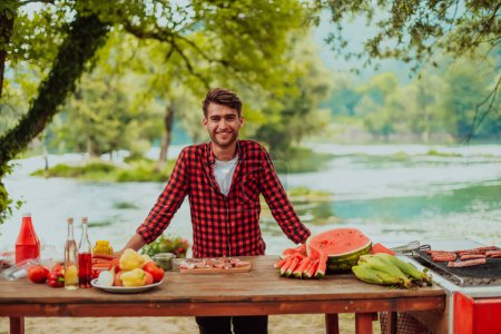 Photo for A man preparing a delicious dinner for his friends who are having fun by the river in nature. - Royalty Free Image