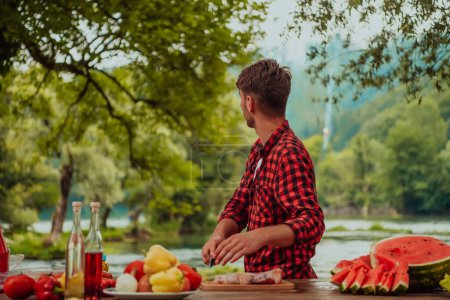 Téléchargez les photos : A man preparing a delicious dinner for his friends who are having fun by the river in nature. - en image libre de droit