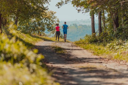 Téléchargez les photos : Couple enjoying in a healthy lifestyle while jogging on a country road through the beautiful sunny forest, exercise and fitness concept. - en image libre de droit