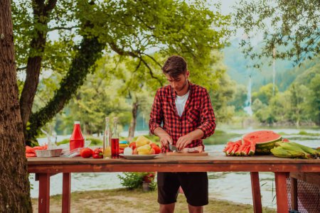 Foto de A man preparing a delicious dinner for his friends who are having fun by the river in nature. - Imagen libre de derechos