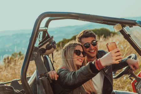 Foto de Joven feliz pareja emocionada disfrutando de hermoso día soleado tomando foto selfie mientras conduce un coche todoterreno buggy en la naturaleza de montaña. - Imagen libre de derechos