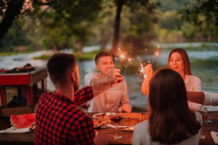 Téléchargez les photos : Groupe d'amis heureux célébrant vacances en utilisant des arroseurs et boire du vin rouge tout en ayant pique-nique dîner français en plein air près de la rivière lors d'une belle soirée d'été dans la nature. - en image libre de droit