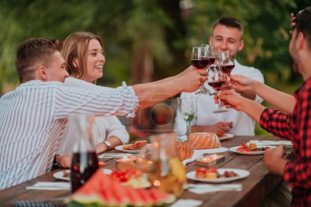 Téléchargez les photos : Groupe d'amis heureux toasting verre de vin rouge tout en ayant pique-nique dîner français en plein air pendant les vacances d'été près de la rivière à la belle nature. - en image libre de droit
