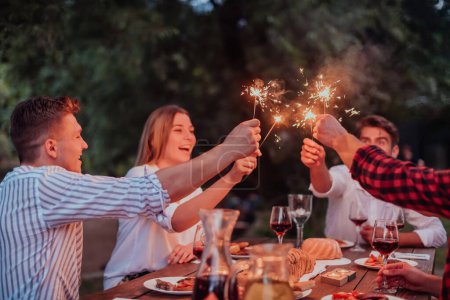 Téléchargez les photos : Groupe d'amis heureux célébrant vacances en utilisant des arroseurs et boire du vin rouge tout en ayant pique-nique dîner français en plein air près de la rivière lors d'une belle soirée d'été dans la nature. - en image libre de droit