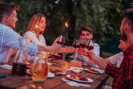 Foto de Grupo de amigos felices celebrando vacaciones de vacaciones con aspersores y beber vino tinto durante el picnic cena francesa al aire libre cerca del río en la hermosa noche de verano en la naturaleza
. - Imagen libre de derechos