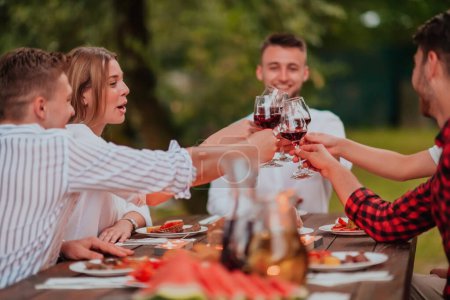 Téléchargez les photos : Groupe d'amis heureux toasting verre de vin rouge tout en ayant pique-nique dîner français en plein air pendant les vacances d'été près de la rivière à la belle nature. - en image libre de droit