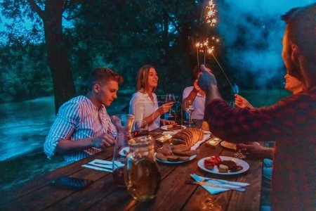 Foto de Grupo de amigos felices celebrando vacaciones de vacaciones con aspersores y beber vino tinto durante el picnic cena francesa al aire libre cerca del río en la hermosa noche de verano en la naturaleza
. - Imagen libre de derechos