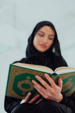 Téléchargez les photos : Jeune femme musulmane traditionnelle lisant le Coran sur le canapé avant le dîner iftar lors d'un festin du Ramadan à la maison. - en image libre de droit
